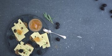 Glasteller mit 3 Brotscheiben belegt mit Allgäuer Emmentaler, Konfitüre und Brombeeren, daneben ein Löffel