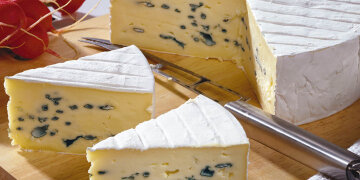 2 Tortenstücke Blauschimmelkäse auf einem Holzbrett, dahinter ein Käsemesser und ein Laib
