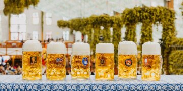 6 Maßkrüge der Münchener Oktoberfestbier-Brauereien, im Hintergrund Festzeltbetrieb und Hopfendeko