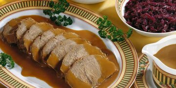 8 Scheiben Sauerbraten in Soße auf einer Servierplatte, daneben Rotkohl und Soße in Schüsseln.