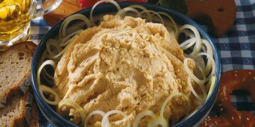 Obazda in einer Keramikschüssel, mit Zwiebelringen garniert, dazu Brotscheiben, eine Breze und ein Krug Bier