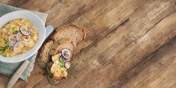 eine weiße Schüssel mit Obazda, dekoriert mit Zweibelringen und Schnittlauch, daneben ein Messer und 2 Scheiben Brot, eins davon bestrichen