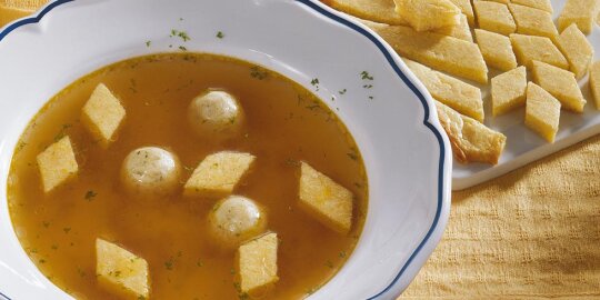 ein Teller Hochzeitssuppe, mit 5 Biskuitrauten und 3 Markklöschen, daneben ein Brettchen mit geschnittenem Biskuit
