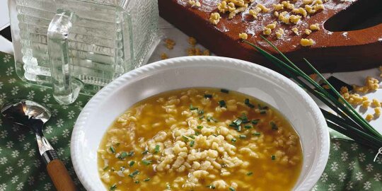 ein Teller Riebelesuppe, daneben ein Löffel, Schnittlauchbund sowie ein Brettchen mit weiteren Riebele