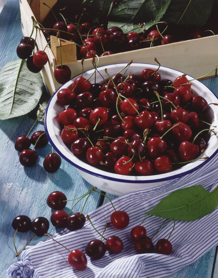 eine Schale mit Süßkirschen auf einem Tisch neben einer Obstkiste mit weiteren