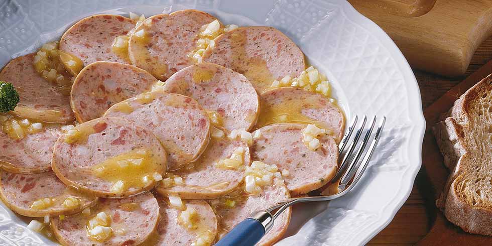Mehrere Wurtscheiben, mit Essig-Öl-Marinade und Zwiebeln auf einem Teller, daneben eine Gabel und eine Scheibe Brot