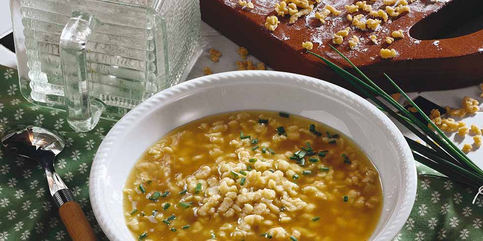 ein Teller Riebelesuppe, daneben ein Löffel, Schnittlauchbund sowie ein Brettchen mit weiteren Riebele